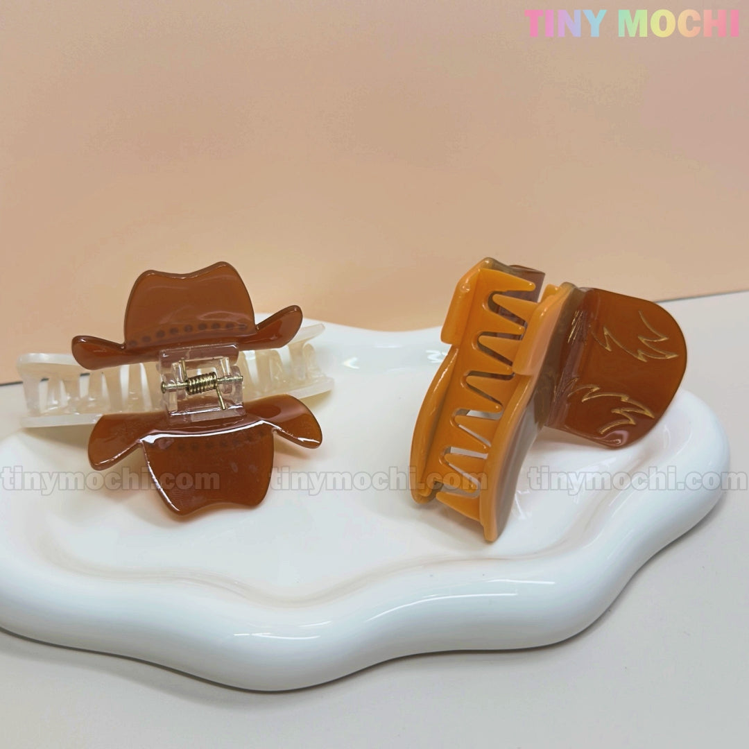 Two Tiny Mochi Western Cowboy Hair Claw Clips with boot and hat design rest on a white tray—one shows the clasp, the other displays cute cowgirl details. The background is light pink.