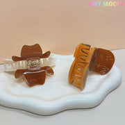 Two Tiny Mochi Western Cowboy Hair Claw Clips with boot and hat design rest on a white tray—one shows the clasp, the other displays cute cowgirl details. The background is light pink.