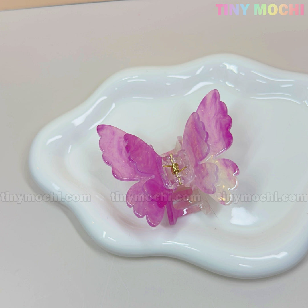 A NEW UV Color-Changing Butterfly Hair Claw Clip by Tiny Mochi sits on a white, wavy-edged ceramic dish. This double layer acetate pastel accessory features a magical spring vibe and pastel Fairycore aesthetic, with the TINY MOCHI watermark visible.