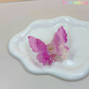 A NEW UV Color-Changing Butterfly Hair Claw Clip by Tiny Mochi sits on a white, wavy-edged ceramic dish. This double layer acetate pastel accessory features a magical spring vibe and pastel Fairycore aesthetic, with the TINY MOCHI watermark visible.