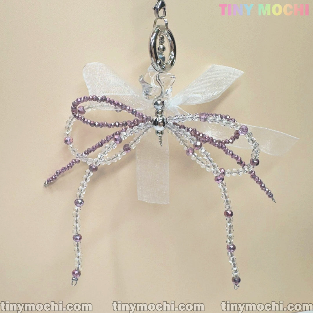 The Delicate Beaded Bow Charm by Tiny Mochi features purple and clear beads, a sheer white ribbon bow, and a metal loop. This handmade Y2K & coquette keychain is shown on a light beige background with TINY MOCHI in colorful text at top right.