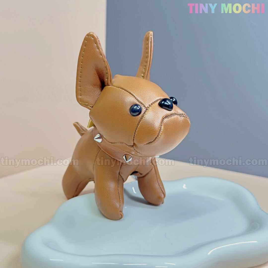 A small, brown French Bulldog Keychain Coin Purse by Tiny Mochi stands on a pale blue tray, featuring faux leather, big ears, black button eyes, and a studded collar—an adorable mini zipper pouch for dog lovers. Soft pastels frame the Tiny Mochi logo.