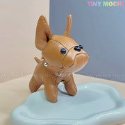 A small, brown French Bulldog Keychain Coin Purse by Tiny Mochi stands on a pale blue tray, featuring faux leather, big ears, black button eyes, and a studded collar—an adorable mini zipper pouch for dog lovers. Soft pastels frame the Tiny Mochi logo.