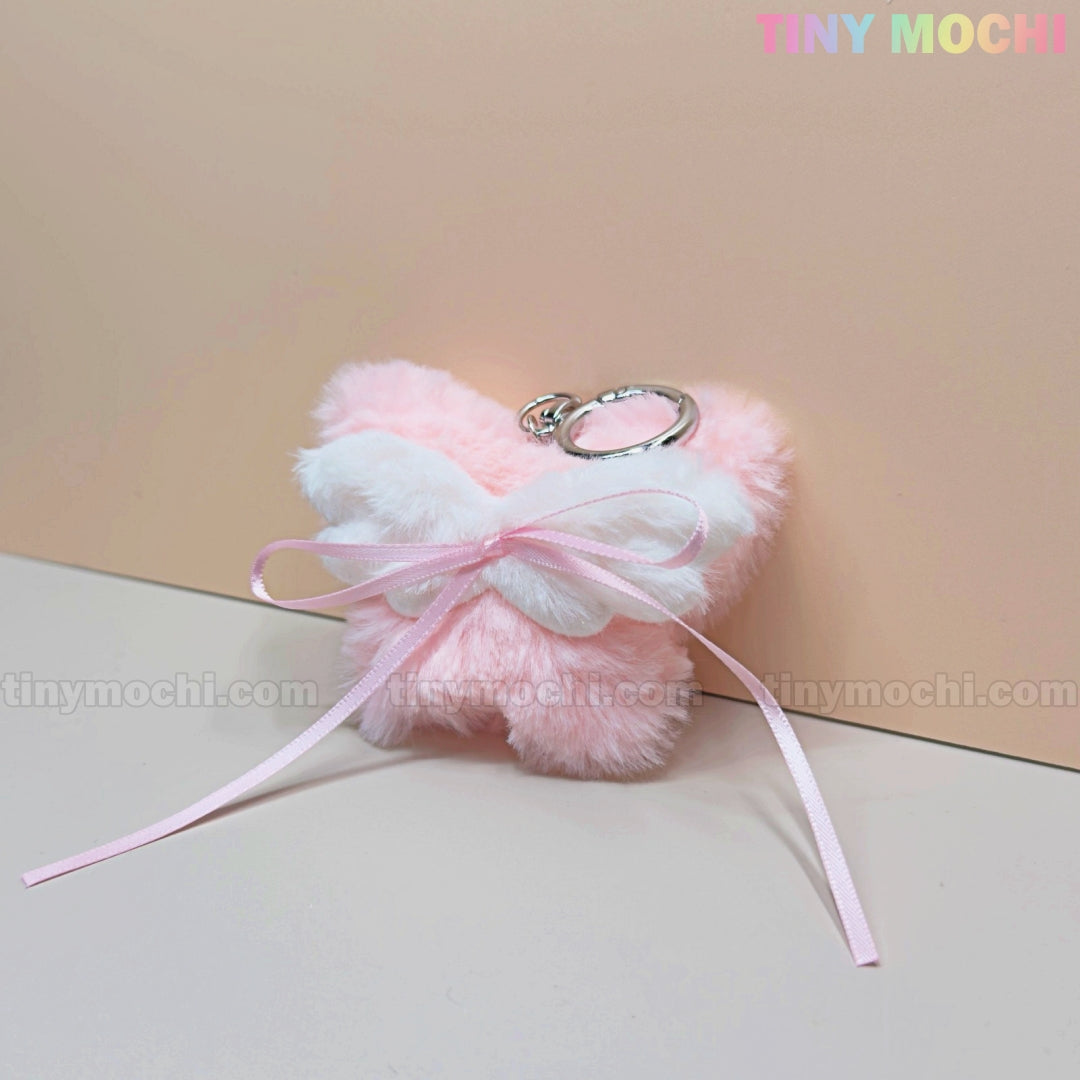 The Tiny Mochi Plush Butterfly Bow Keychain is a pink fuzzy bag charm with white wings and a silver keyring—perfect as a soft girl or coquette cute gift and accessory, shown on a light pink background.