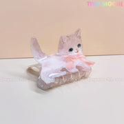 A Kawaii hair claw clip shaped like a light brown cat with blue eyes and a pink bow, set on a pale beige background. Tiny Mochis pastel logo appears at the top right—perfect for fans of cute pet-themed hair accessories!.