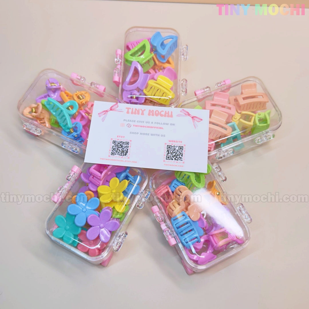Five clear boxes, each holding the Tiny Mochi Kids Hair Clips Set with 10 colorful kawaii claw clips in butterfly, flower & heart designs, are arranged in a star around a Tiny Mochi card. The cute, kid-safe clips come in pastel shades.