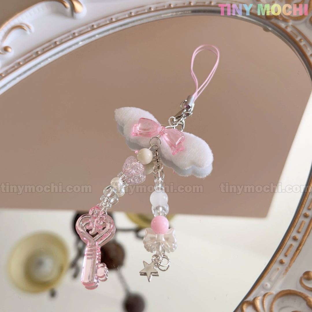 The Kawaii Angel Wing Keychain – Pink Heart Charm by Tiny Mochi features plush bows, crystal-like beads, a pink heart charm, and star charms on a pink string—a perfect soft girl accessory reflected in an ornate mirror.
