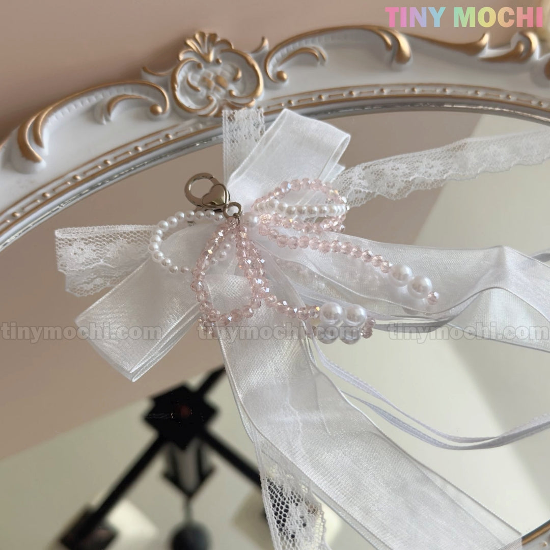 A delicate Lace Bow Ribbon Keychain – Pearl Angelcore Bag Charm Clip by Tiny Mochi, featuring white lace, organza bow, and pink and white pearl beads, is clipped to an ornate mirror with the Tiny Mochi brand in the top right corner.