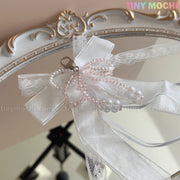 A delicate Lace Bow Ribbon Keychain – Pearl Angelcore Bag Charm Clip by Tiny Mochi, featuring white lace, organza bow, and pink and white pearl beads, is clipped to an ornate mirror with the Tiny Mochi brand in the top right corner.