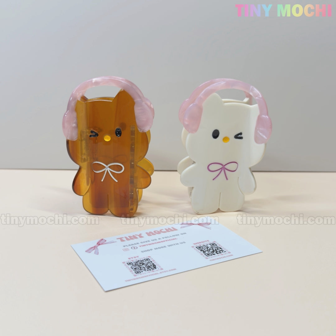 Two Kawaii Bunny & Bear Hair Claw Clips with pink headphones by Tiny Mochi, one amber and one white, stand upright on a beige surface with a business card in front. The Tiny Mochi logo is visible in the top right corner.