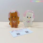 Two Kawaii Bunny & Bear Hair Claw Clips with pink headphones by Tiny Mochi, one amber and one white, stand upright on a beige surface with a business card in front. The Tiny Mochi logo is visible in the top right corner.