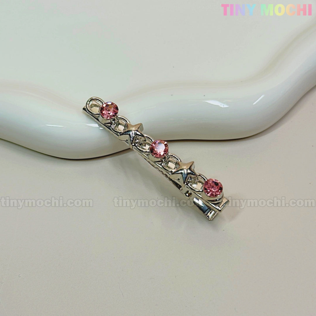 A silver hair clip with three pink gems and two silver stars rests on a cream ceramic dish. Ideal for Kawaii Gem Hair Clips for Girls by Tiny Mochi, these sparkly Y2K pins add Coquette flair. Tiny Mochi appears in rainbow text at the top right corner.