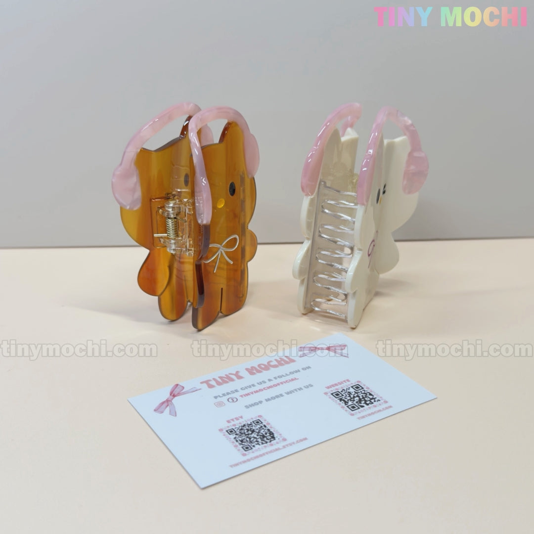 Two Kawaii Bunny & Bear Hair Claw Clips with Pink Headphones by Tiny Mochi stand upright on a beige surface. A business card with QR codes and branding lies in front, making these the perfect cute accessory for any look.