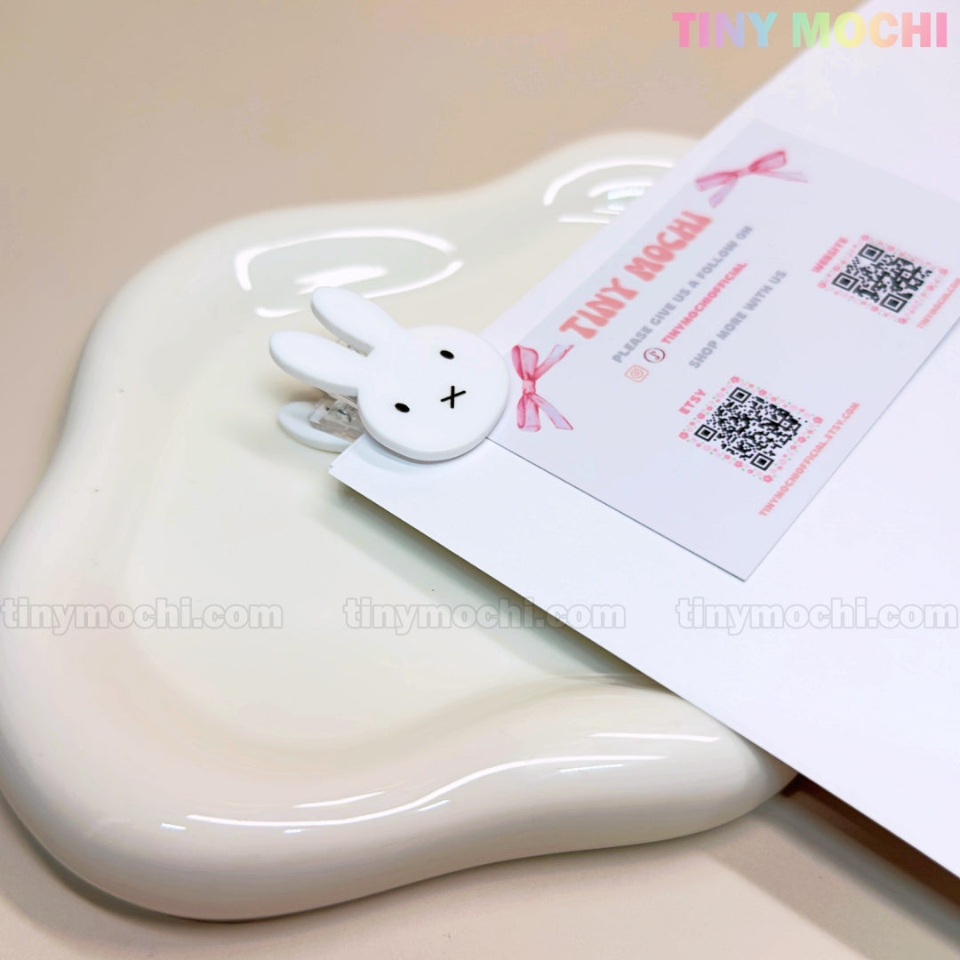 A Kawaii Bunny Paper Clip from Tiny Mochi, inspired by Miffy and perfect for desk decor, holds a business card atop a glossy white ceramic dish—combining cute organization with adorable character stationery. The card features QR codes.