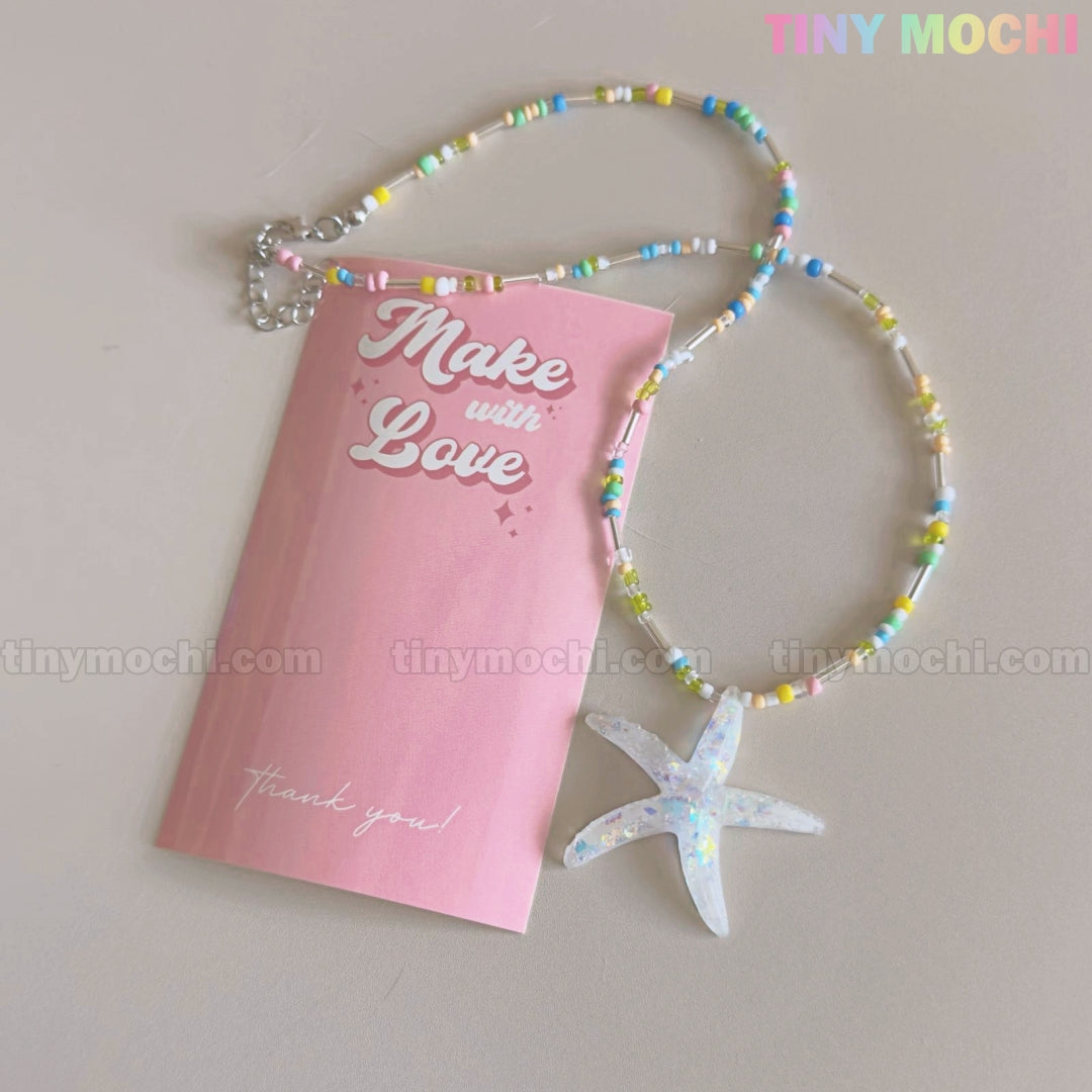 The Tiny Mochi Starfish Beach Necklace features an iridescent blue starfish pendant on a beaded choker, displayed beside a pink Make with Love card—creating a playful summer vibe and the perfect ocean-inspired gift for girls and teens.