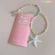 The Tiny Mochi Starfish Beach Necklace features an iridescent blue starfish pendant on a beaded choker, displayed beside a pink Make with Love card—creating a playful summer vibe and the perfect ocean-inspired gift for girls and teens.