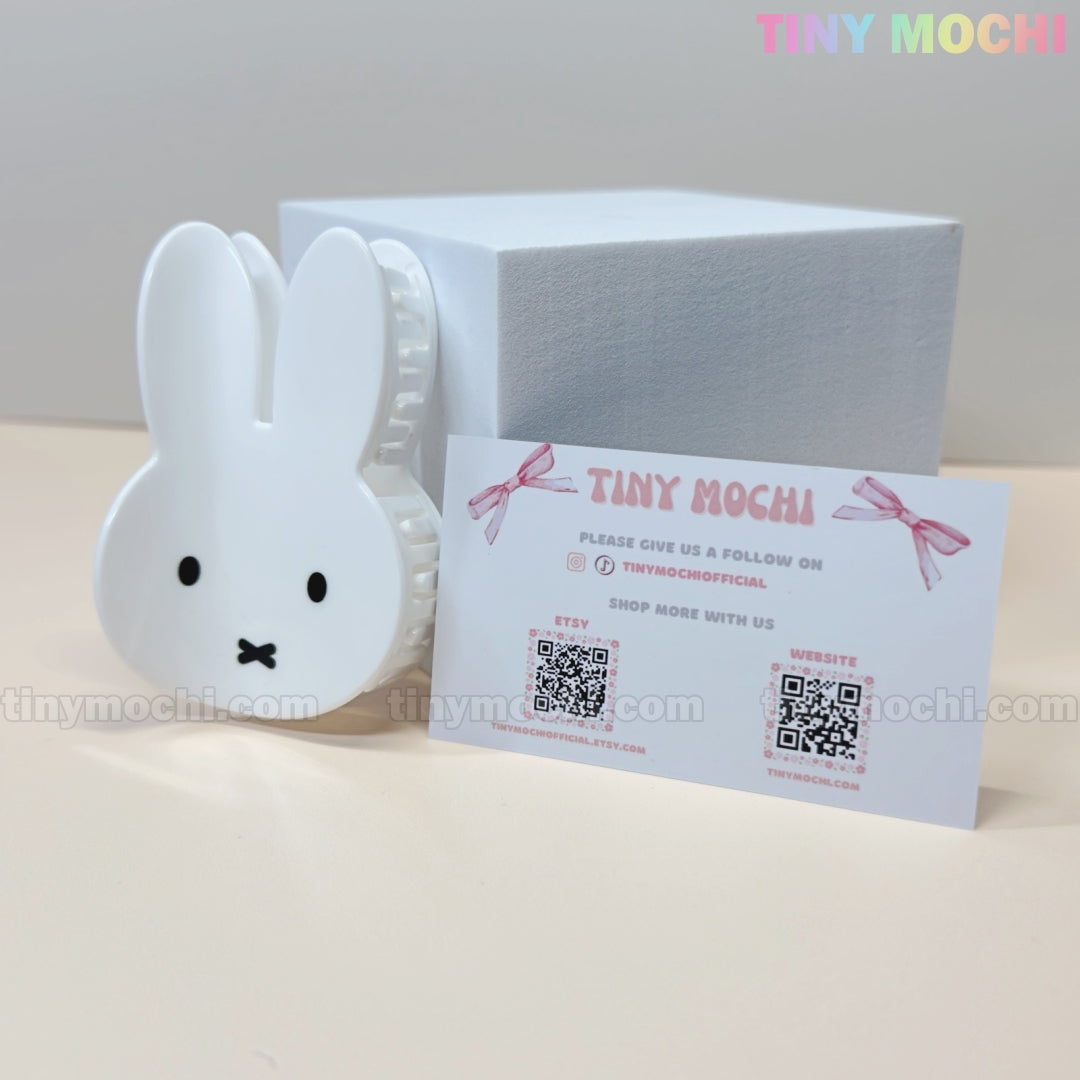 A white Kawaii Bunny Face Hair Clip – White Rabbit Claw Clip by Tiny Mochi, a branded business card with QR codes and contact info, and a white box are arranged on a cream surface—ideal for fans of cute hair accessories.