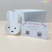 A white Kawaii Bunny Face Hair Clip – White Rabbit Claw Clip by Tiny Mochi, a branded business card with QR codes and contact info, and a white box are arranged on a cream surface—ideal for fans of cute hair accessories.