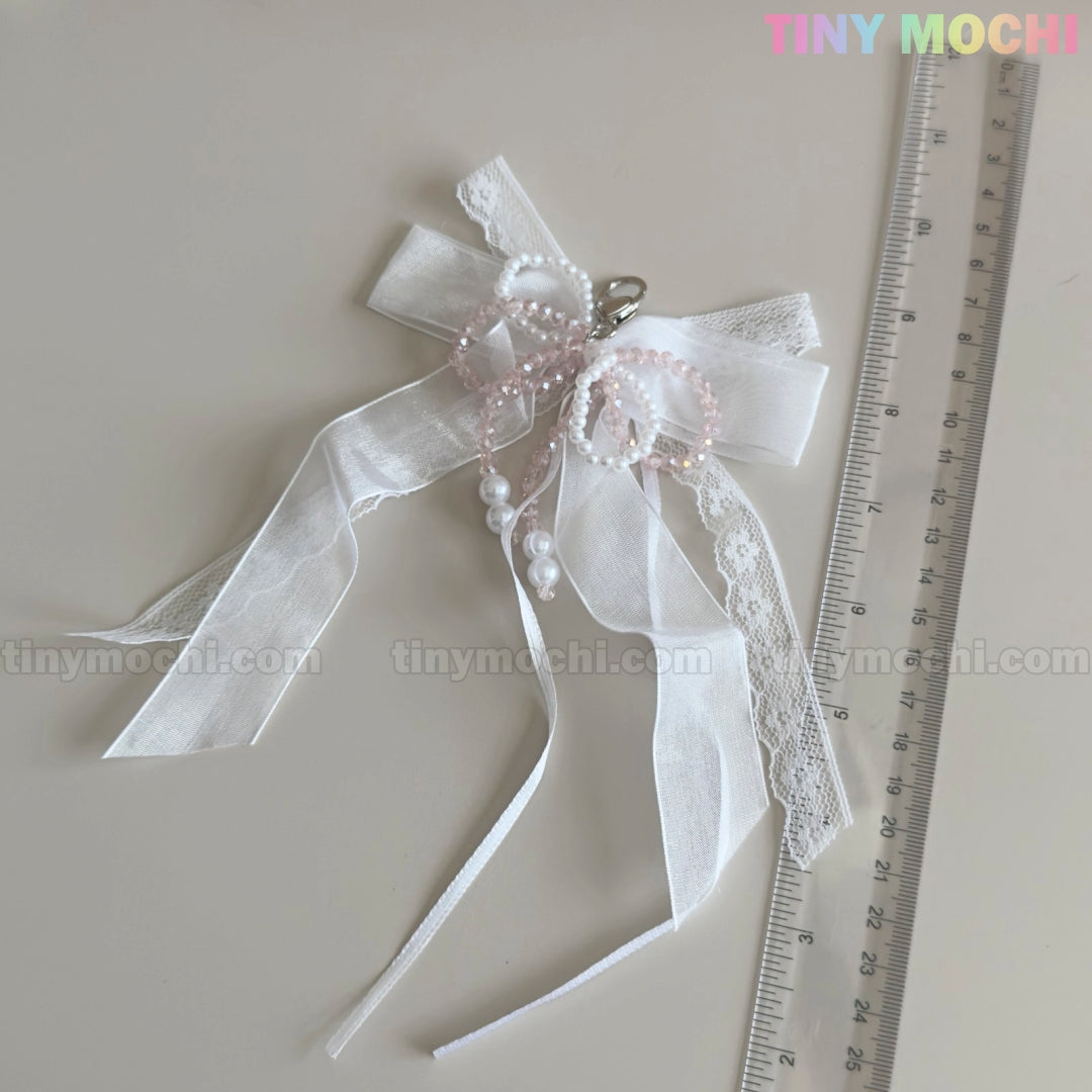 The Tiny Mochi Lace Bow Ribbon Keychain – Pearl Angelcore Bag Charm Clip, featuring white lace, pearls, and pink beads, is shown by a transparent ruler on a light surface. The pastel Tiny Mochi logo appears at the top right.