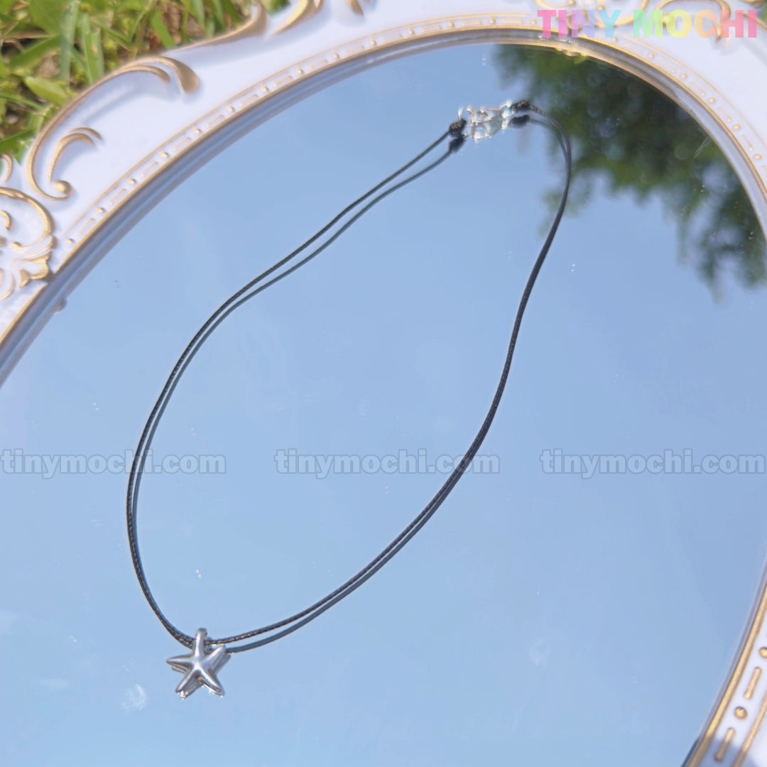 The Tiny Mochi Sterling Silver Starfish Necklace on a black cord, a minimalist ocean jewelry piece and handmade gift for him or her, is displayed on an ornate mirror reflecting greenery and sky with tinymochi.com branding visible.