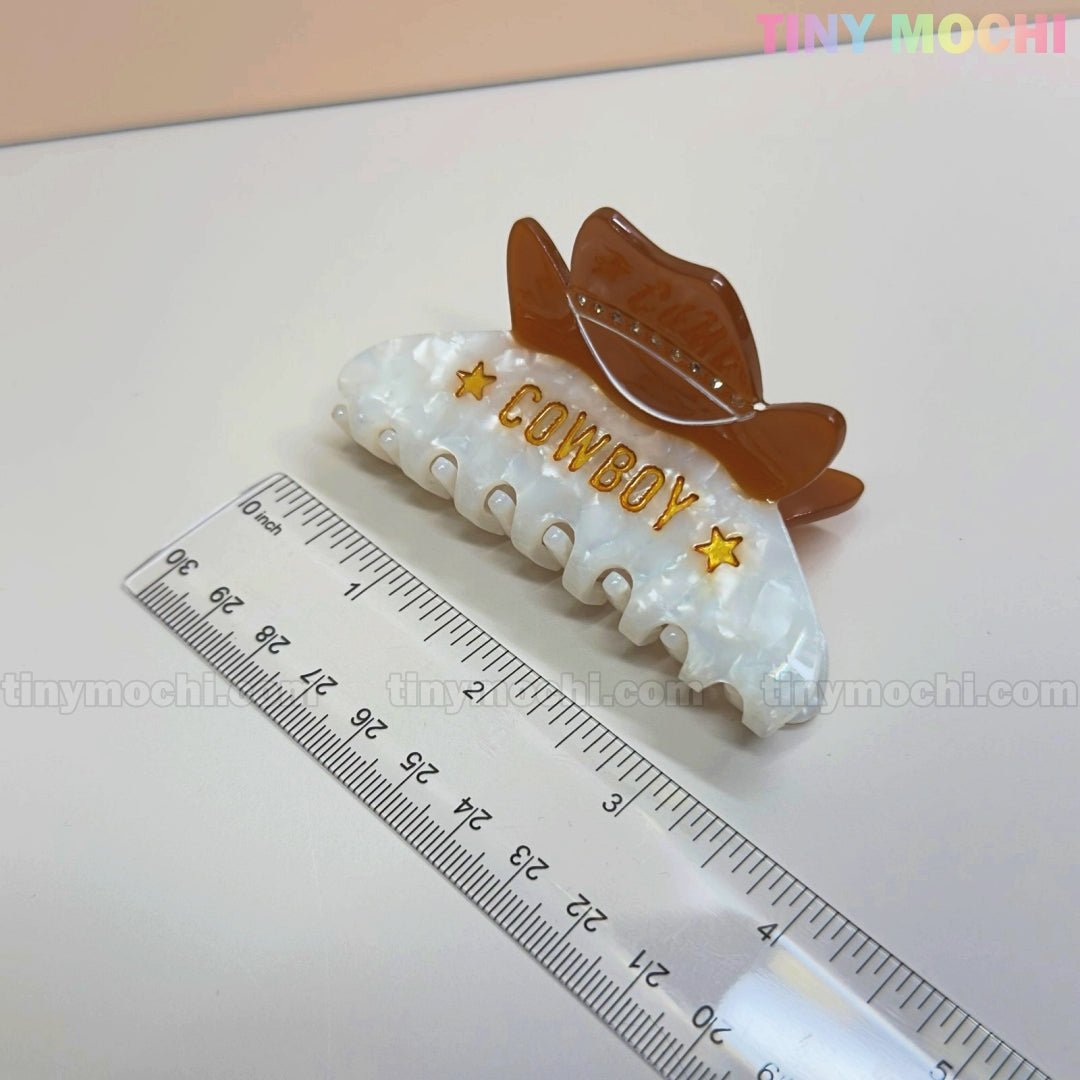 A Tiny Mochi Western Cowboy Hair Claw Clip for women and girls, featuring a cute white and brown cowgirl hat with gold COWBOY and star accents, is shown beside a clear ruler. Perfect Western accessory for hair styling fans.