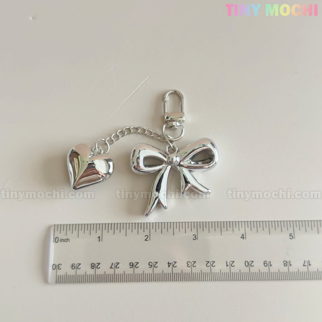 A Silver Bow Heart Keychain – Kawaii Mirror Bag Charm for Backpacks by Tiny Mochi is shown next to a ruler, measuring about 7 cm long. TINYMOSCHI.COM and Tiny Mochi are watermarked on the white background.