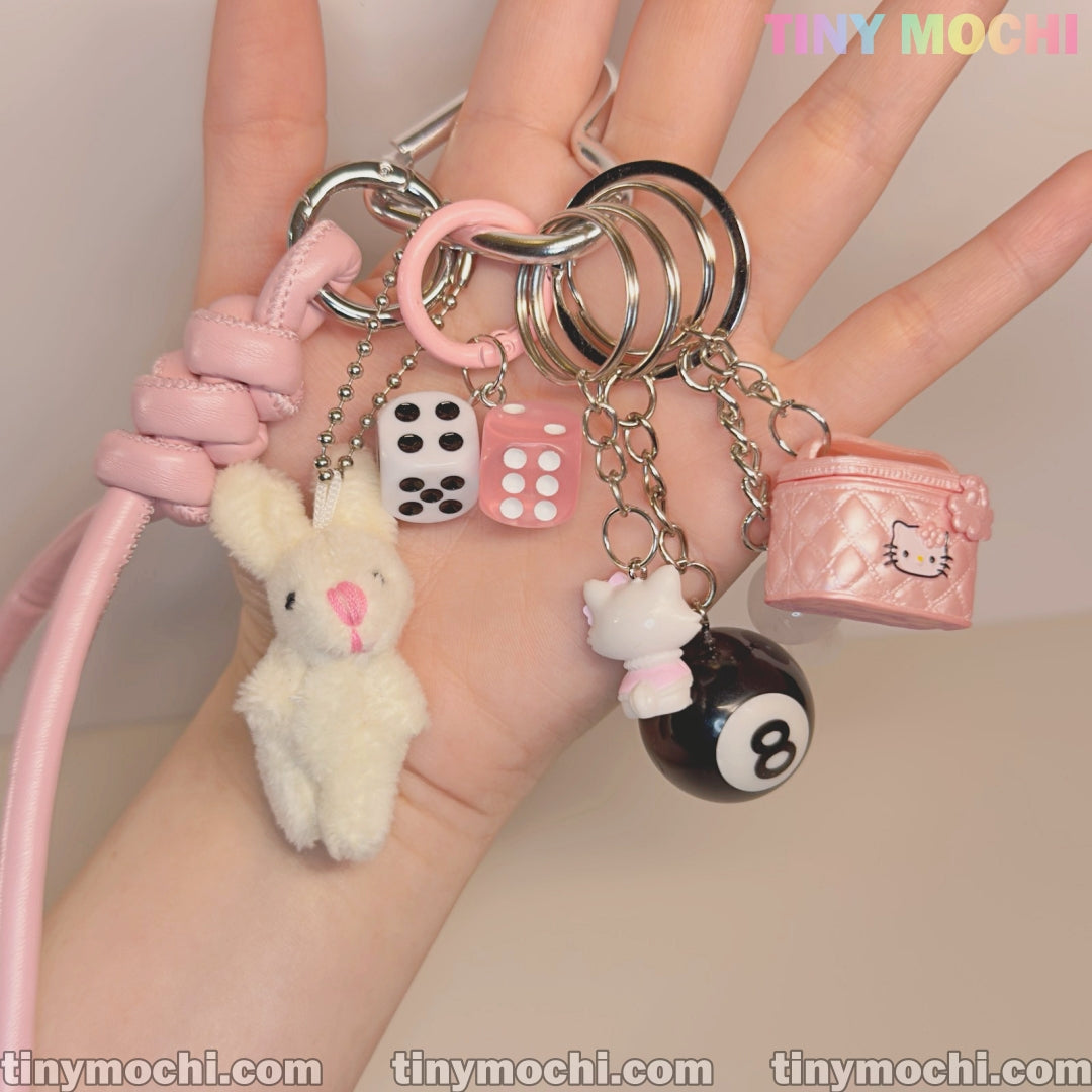 A hand holds the Cute Bunny and Charm Keychain by Tiny Mochi, featuring a white plush bunny, black 8-ball, pink dice, Hello Kitty figure, and pink quilted purse charm—an aesthetic accessory for purses and backpacks.