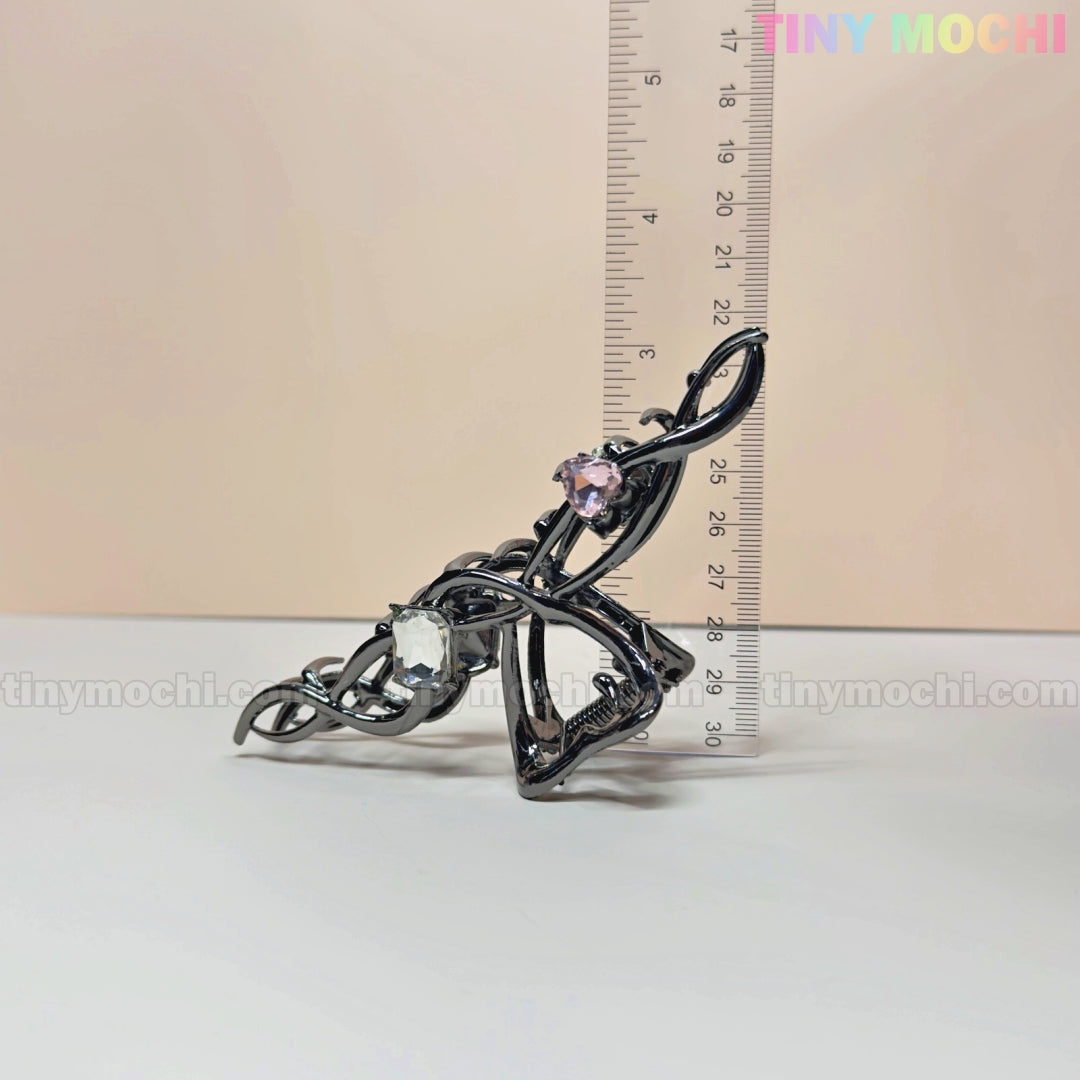 The Tiny Mochi Dark Silver Crystal Heart Hair Claw is an elegant metal clip for women, featuring a strong grip and two gemstones—one pink, one clear—plus a swirling design. Shown next to a ruler, it measures about 9 cm tall.
