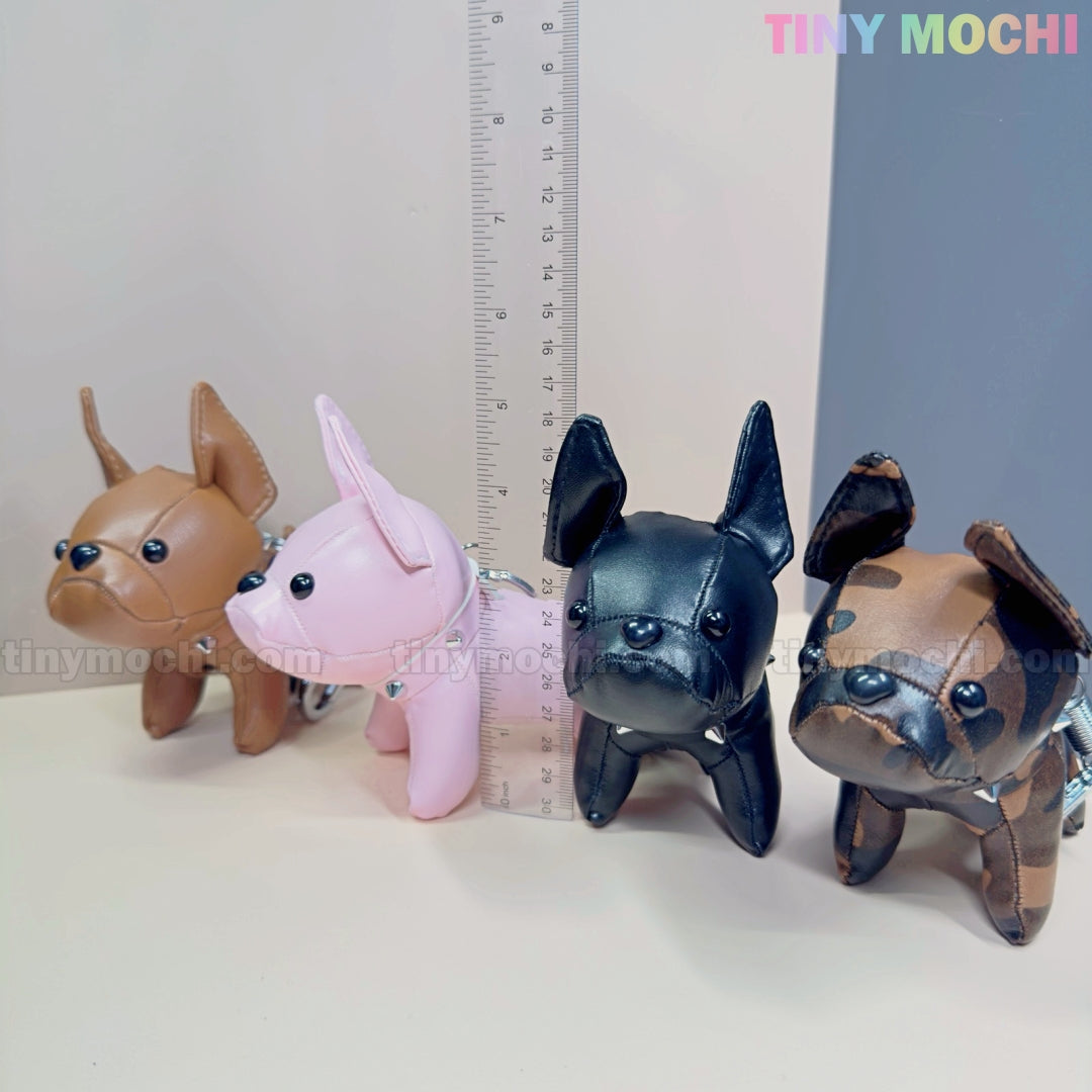 Four mini French Bulldog keychain coin purses by Tiny Mochi in brown, pink, black, and brown-black are lined up beside a clear ruler on a light surface. Cute faux leather puppy bag charms for dog lovers. Logo displayed at top right corner.