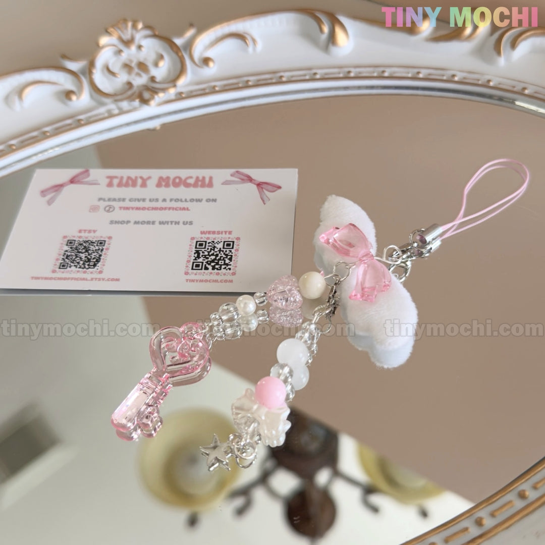 The Kawaii Angel Wing Keychain – Pink Heart Charm Bag Accessory by Tiny Mochi rests on a mirror alongside a business card, its angelcore charm and pink accents reflected in the ornate frame—perfect for adding cute, soft girl vibes.