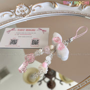 The Kawaii Angel Wing Keychain – Pink Heart Charm Bag Accessory by Tiny Mochi rests on a mirror alongside a business card, its angelcore charm and pink accents reflected in the ornate frame—perfect for adding cute, soft girl vibes.