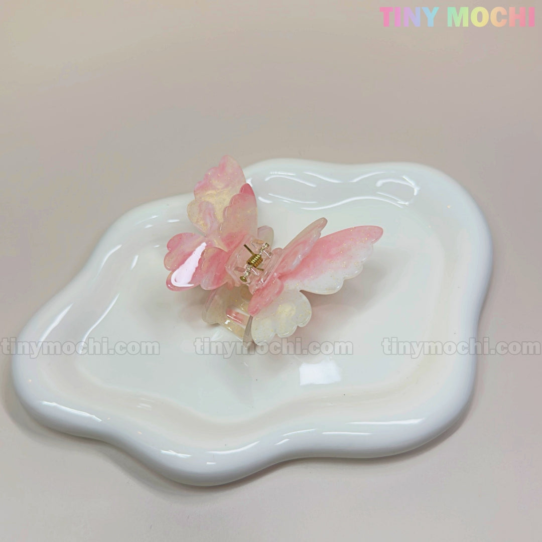 A NEW UV Color-Changing Butterfly Hair Claw Clip by Tiny Mochi, featuring double-layer acetate in pastel hues, rests on a wavy white ceramic dish. The image and pastel Tiny Mochi logo evoke a dreamy Fairycore vibe.