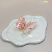 A NEW UV Color-Changing Butterfly Hair Claw Clip by Tiny Mochi, featuring double-layer acetate in pastel hues, rests on a wavy white ceramic dish. The image and pastel Tiny Mochi logo evoke a dreamy Fairycore vibe.