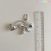 The Silver Bow Heart Keychain – Kawaii Mirror Bag Charm for Backpacks by Tiny Mochi adds a coquette touch next to a clear ruler on a white surface, with the Tiny Mochi logo in the top right corner.