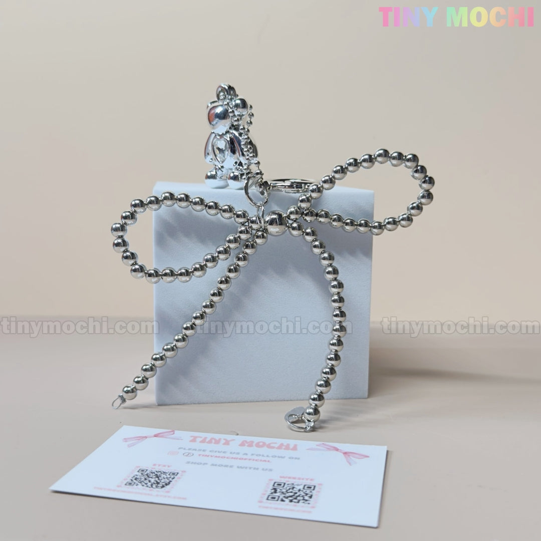 A small white gift box with a silver beaded bow is topped with the Silver Teddy Bear Bow Keychain by Tiny Mochi, perfect as a cute bag charm for women, teens & kawaii lovers. Includes a business card with TINY MOCHI and QR codes.