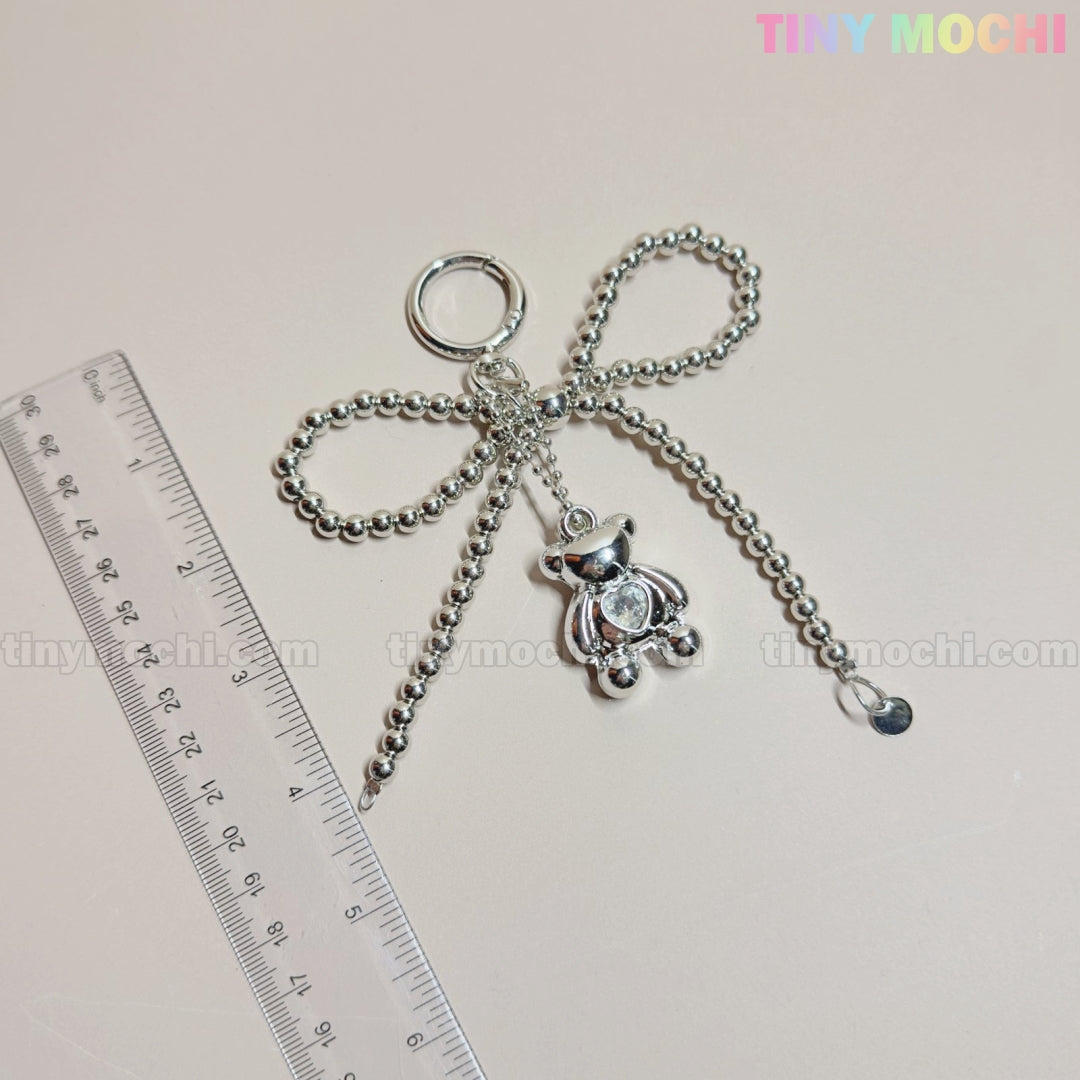 The Silver Teddy Bear Bow Keychain by Tiny Mochi, a cute bag charm accessory for women, teens, and kawaii lovers, hangs beside a clear ruler on a light surface. The branding TINY MOCHI and tinymochi.com are faintly visible.