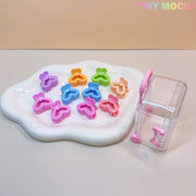A white tray displays the Tiny Mochi Kids Hair Clips Set with 10 colorful kawaii butterfly, flower, and heart-shaped clips. Beside it is a mini travel case resembling a clear gacha capsule machine with a pink handle.