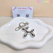 The Balloon Dog Keychain Silver Chrome Bag Charm with Star Accent from Tiny Mochi shines on a wavy white dish, paired with a pastel-branded business card—perfect as a cute metallic purse accessory or gift.