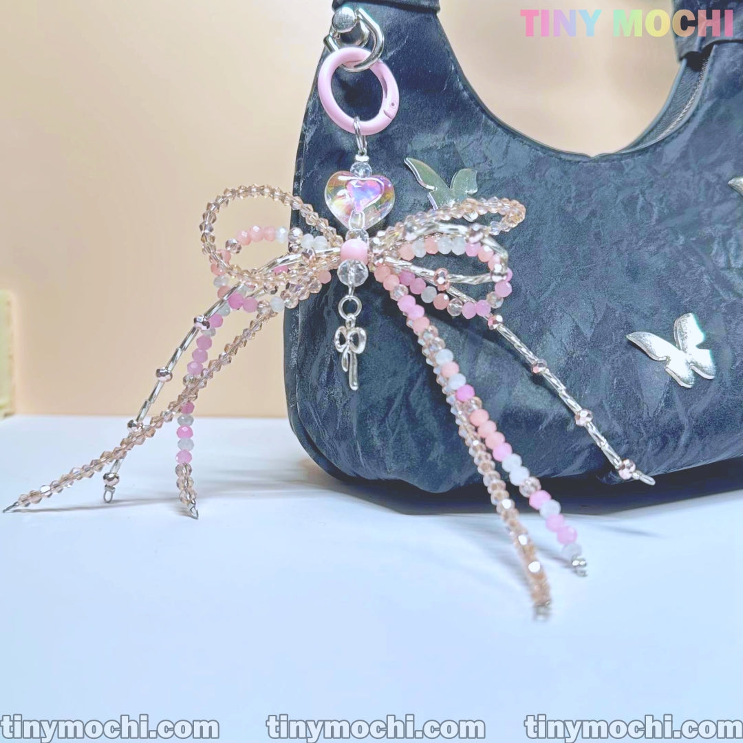 The image shows Tiny Mochi’s Iridescent Beaded Bow Bag Charm, an elegant fairycore keychain for purses or backpacks. It features a pastel bow, iridescent beads, a heart gem, and a key pendant on a pink ring. “TINYMOSCHI” is displayed.