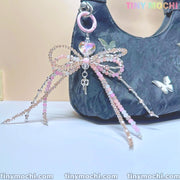 The image shows Tiny Mochi’s Iridescent Beaded Bow Bag Charm, an elegant fairycore keychain for purses or backpacks. It features a pastel bow, iridescent beads, a heart gem, and a key pendant on a pink ring. “TINYMOSCHI” is displayed.