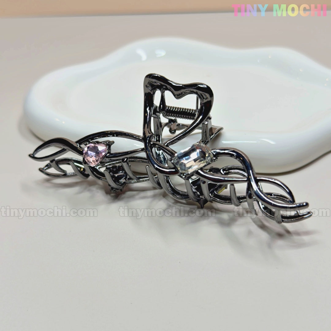 The Dark Silver Crystal Heart Hair Claw by Tiny Mochi features an elegant twisted vine design with pink and clear crystal hearts. Shown on a white dish, this metal clip offers a strong grip. Tiny Mochi’s logo appears in the top right corner.