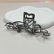 The Dark Silver Crystal Heart Hair Claw by Tiny Mochi features an elegant twisted vine design with pink and clear crystal hearts. Shown on a white dish, this metal clip offers a strong grip. Tiny Mochi’s logo appears in the top right corner.