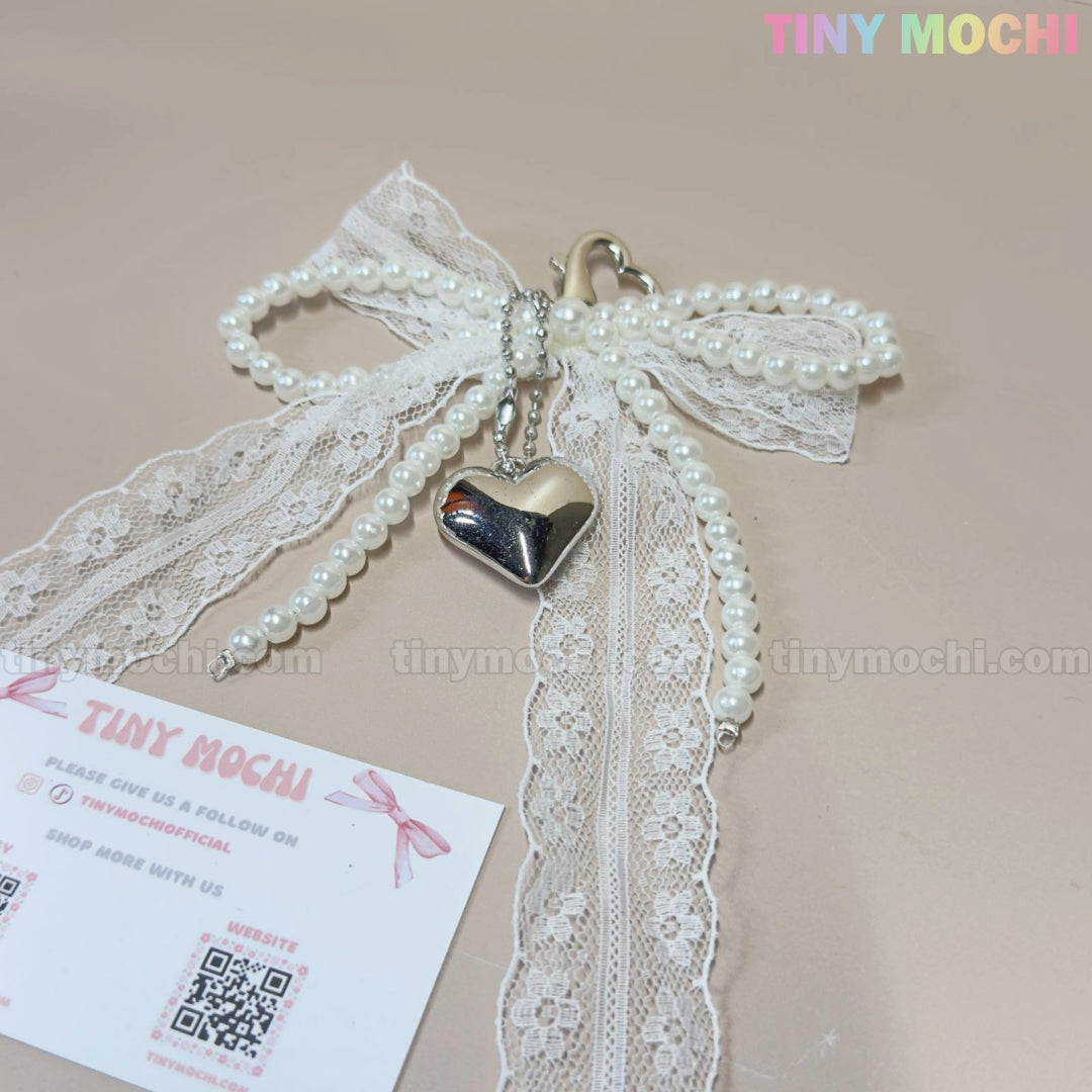 The Pearl Lace Bow Keychain with Silver Heart from Tiny Mochi, perfect for the coquette style, is shown on a beige surface. A Tiny Mochi business card featuring the brands logo is placed nearby.