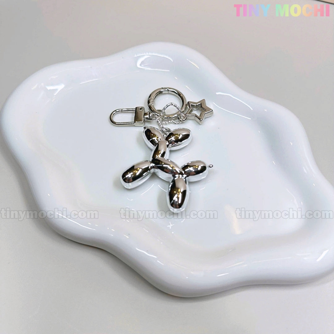 The Balloon Dog Keychain Silver Chrome Bag Charm with Star Accent from Tiny Mochi rests on a wavy white ceramic tray, with the brand name in pastel at the top right, making this metallic purse accessory irresistibly cute.