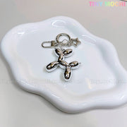 The Balloon Dog Keychain Silver Chrome Bag Charm with Star Accent from Tiny Mochi rests on a wavy white ceramic tray, with the brand name in pastel at the top right, making this metallic purse accessory irresistibly cute.