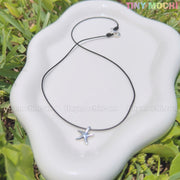 The Tiny Mochi Sterling Silver Starfish Necklace 925 Charm on Black Cord—minimalist ocean jewelry and a handmade gift—is shown on a white wavy dish set on grass, with TINY MOCHI in the top right corner.