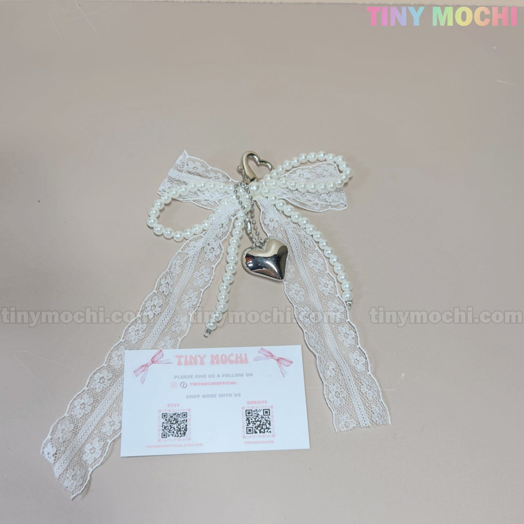 The Pearl Lace Bow Keychain with Silver Heart – Elegant Bag Charm by Tiny Mochi, featuring a coquette design, is displayed above a branded card with QR codes and social media—perfect as a chic accessory for your bag.