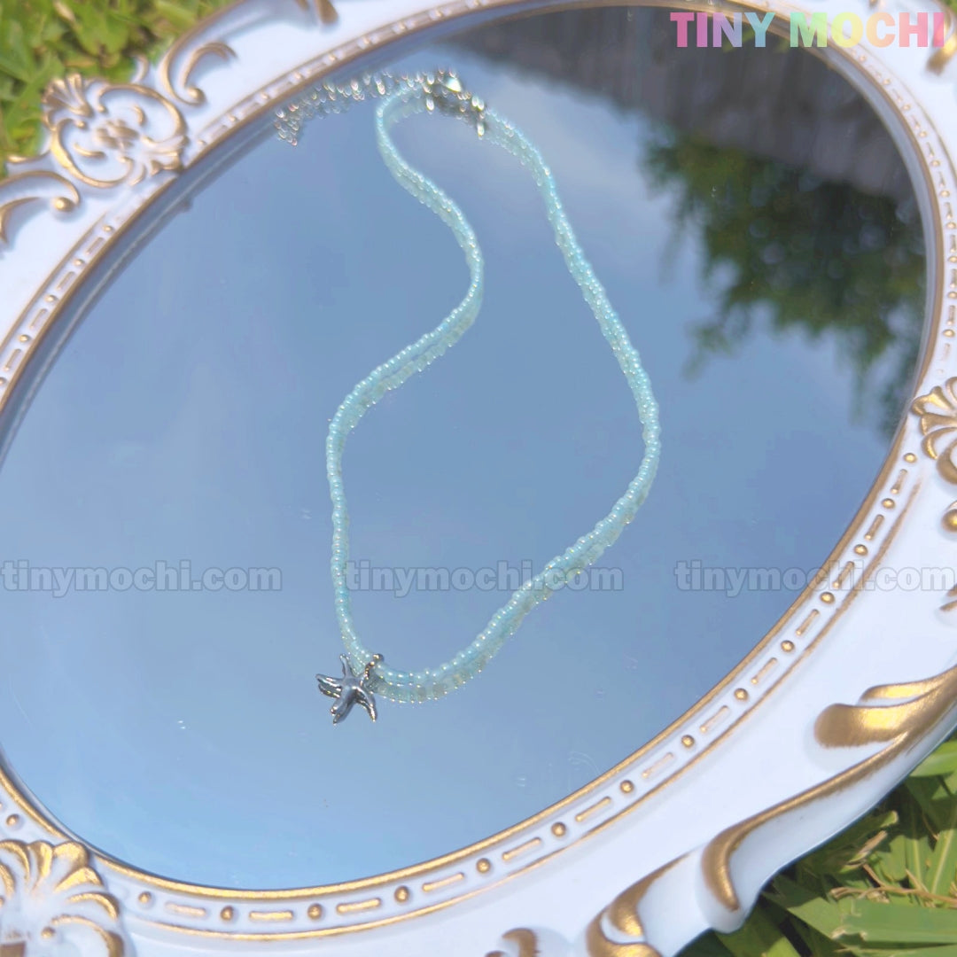 The Silver Starfish Choker Necklace by Tiny Mochi, a dainty light blue beaded beach jewelry piece, is showcased on a decorative mirror reflecting sky and greenery. TINY MOCHI appears in pastel colors at the top right.