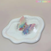 The NEW UV Color-Changing Butterfly Hair Claw Clip by Tiny Mochi rests on a wavy-edged white ceramic dish. A pastel gradient background enhances the Fairycore vibe, with Tiny Mochi in soft colors at the top right.