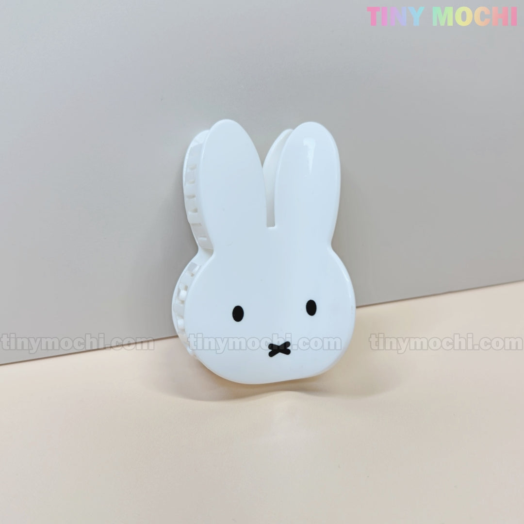 The Kawaii Bunny Face Hair Clip – White Rabbit Claw Clip by Tiny Mochi features a white plastic design shaped like Miffy’s face, with black eyes and an x-mouth, displayed on a beige surface against a pastel Tiny Mochi logo backdrop.